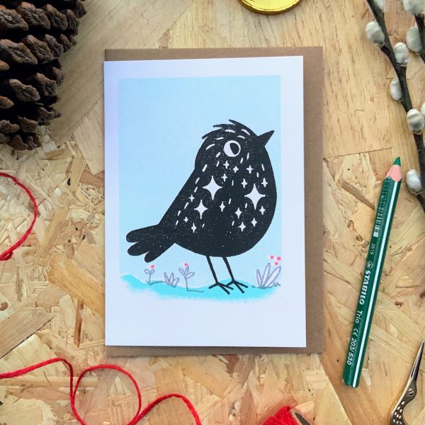 Photograph of a Risograph print greetings card with a black starling bird character standing in the snow.