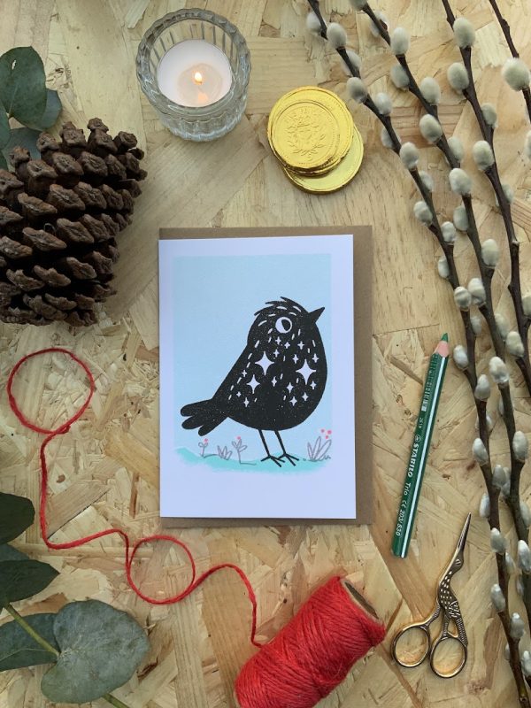 Lay flat photograph with black starling bird character Risograph print card, with red twine, scissors, candle and winter foliage background.